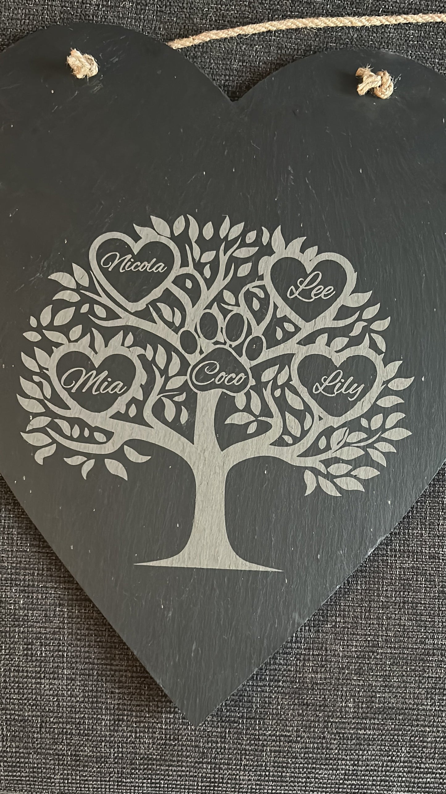 Decorative heart-shaped slate hanging board with tree design and engraved names ina heart on a textured surface.