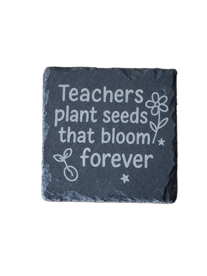 Slate coaster with engraved text 'Teachers plant seeds that bloom forever' on a wooden surface