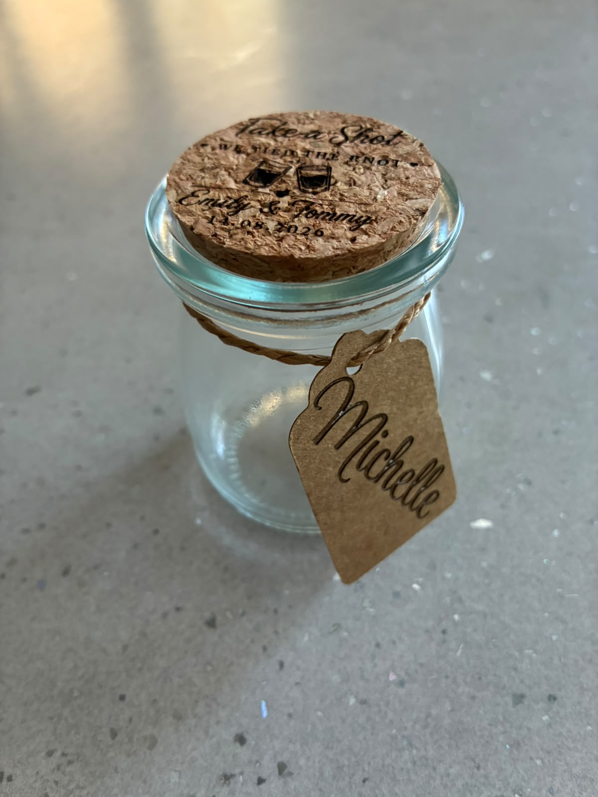 Clear glass wedding shot jar with an engraved cork lid and personalised kraft guest name tag.