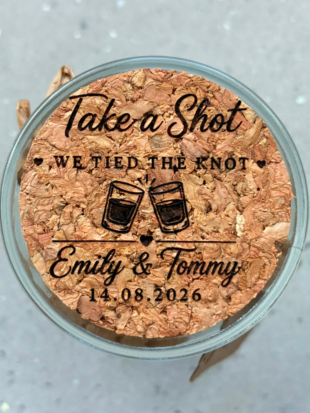 Close-up of a cork jar lid engraved with “Take a Shot, We Tied the Knot”, the couple’s names and wedding date.