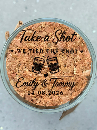Close-up of a cork jar lid engraved with “Take a Shot, We Tied the Knot”, the couple’s names and wedding date.