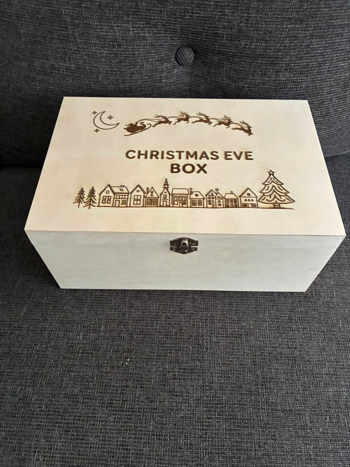 A rectangular wooden Christmas Eve box with engraved holiday illustrations and the text 'CHRISTMAS EVE BOX' on the lid, placed on a dark gray textured fabric surface.
