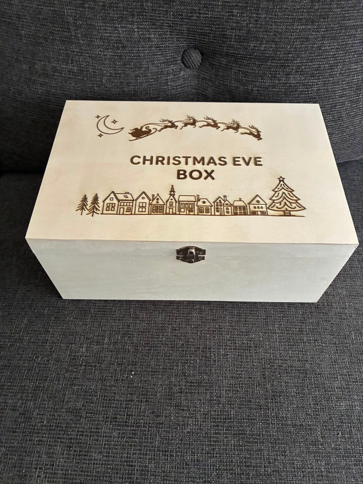 A rectangular wooden Christmas Eve box with engraved holiday illustrations and the text 'CHRISTMAS EVE BOX' on the lid, placed on a dark gray textured fabric surface.