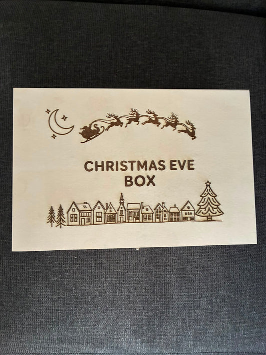 A square cream-colored wooden box lid or insert with laser-engraved Christmas illustrations, including Santa's sleigh, a crescent moon, stars, houses, and a Christmas tree, with the text 'CHRISTMAS EVE BOX' in the center, placed on a dark gray textured fabric background.