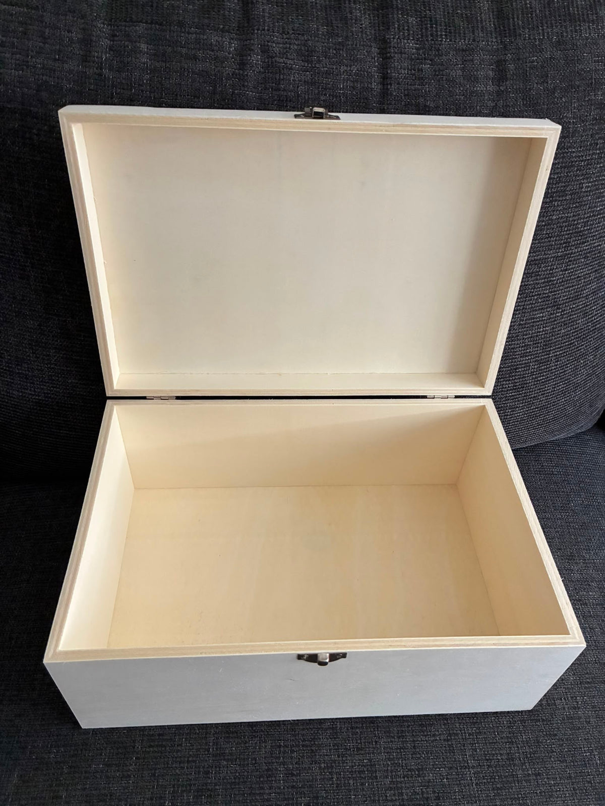 An open light-colored wooden storage box with a hinged lid, photographed against a dark textured fabric background.